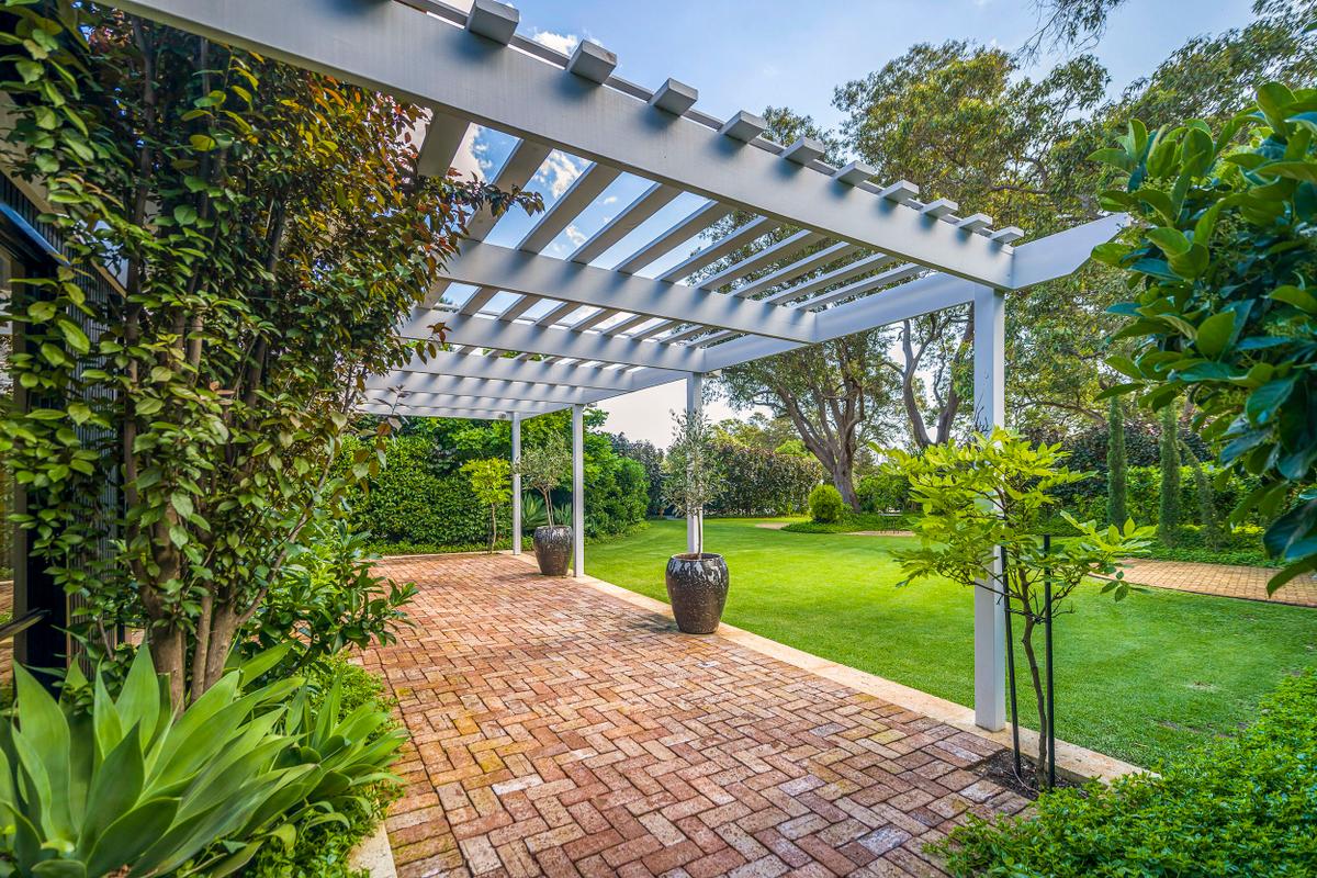 A covered patio with a brick floor, white pergola overhead, and potted plants on the patio in a garden with green grass, trees, and shrubs.